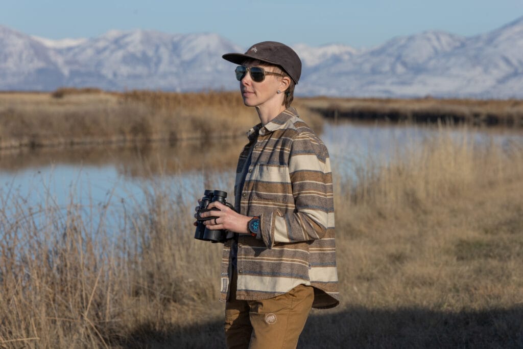 A woman in a Royal Robbins Clouds Rest hemp flannel standing in front of a river and snowy mountains with binocs in hand.