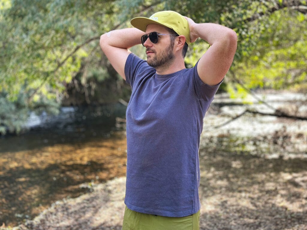 A man stands in the woods wearing a blue Jungmaven Boulder tee.