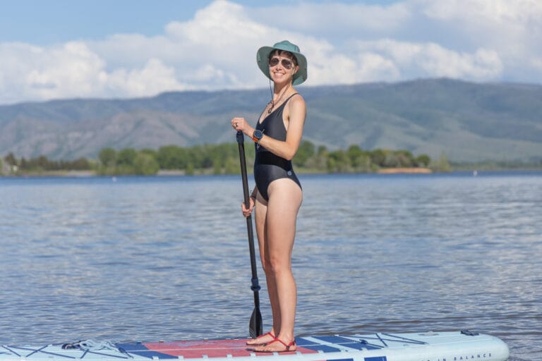 A woman stands on a paddle board in a lake wearing an Outerknown Origin one-piece sustainable swimsuit and a sun hat.