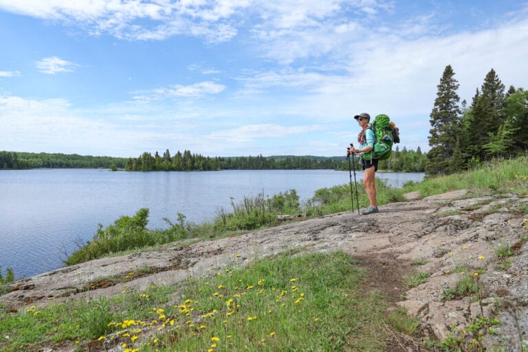 Woman stands on the edge of a lake with a backpack and trekking poles in Isle Royale National Park.