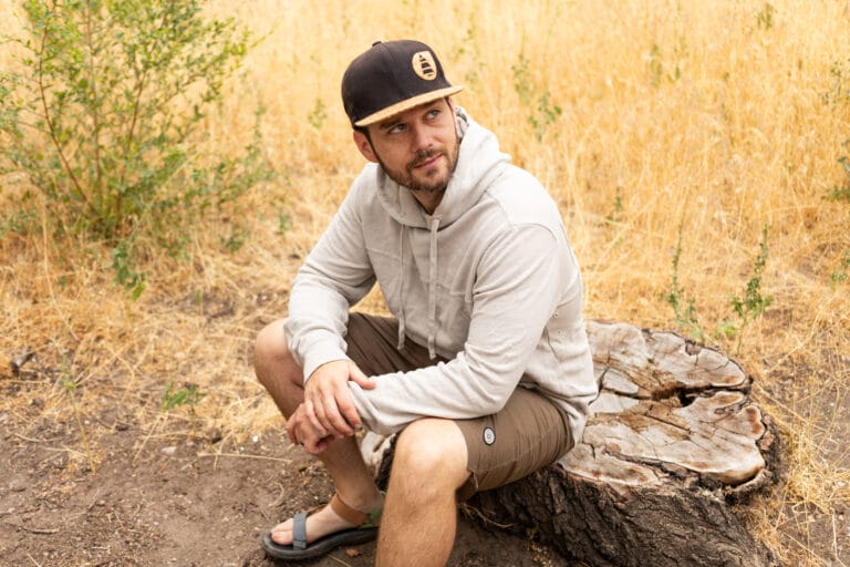 A man sits on a rock outdoors in the Astral Hemp Sunrise Hoodie and Kinisi Shorts.