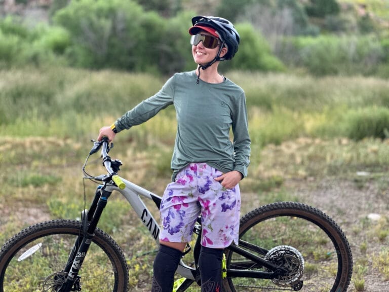 A woman stands by a mountain bike in Wild Rye bike shorts.