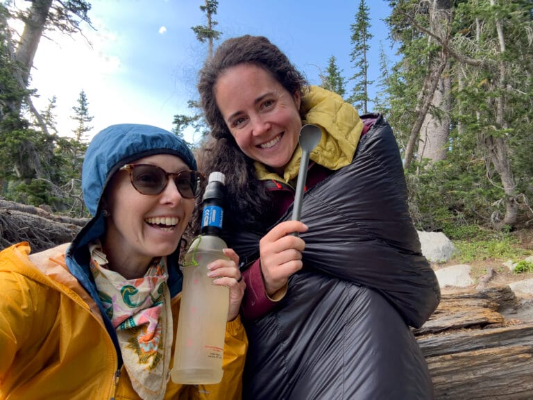 Two women in backpacking gear hold up a spoon and a water bottle with a filter.