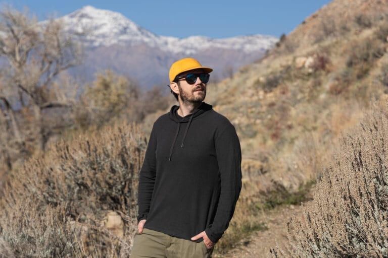 A man stands in front of a mountain on a trail wearing an Astral High Noon hemp hoodie.