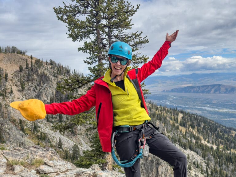 A woman stands in front of mountains excited and arms spread wearing a yellow Houdini ultralight fleece and red rain jacket.