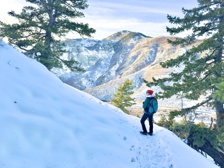 A woman in winter layers stands on a snowy trail with mountains in the background.